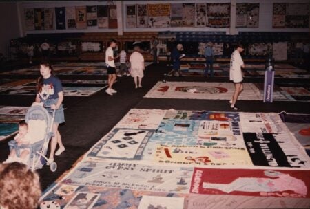 A 1994 photograph of the AIDS Memorial Quilt displayed in a large indoor space in Tampa, Florida. The quilt panels, each dedicated to individuals who lost their lives to AIDS, cover the floor and hang on the walls. People of various ages walk among the quilts, observing and reflecting. A woman pushes a baby stroller, while others read the names and messages stitched onto the panels. A donation station stands in the background.