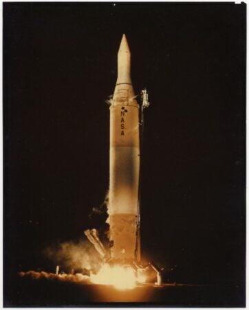 A nighttime photograph of the Juno II rocket launch by NASA. The rocket stands vertically, illuminated by flames and smoke at its base as it lifts off the ground. The NASA logo is visible on the rocket's body, and the dark sky forms the background.