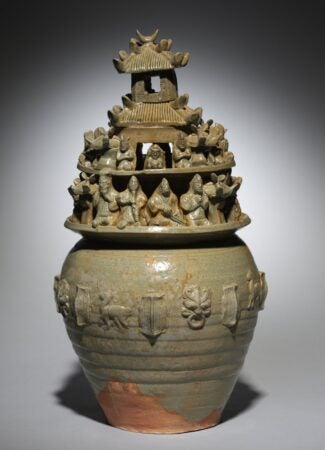 A green-glazed stoneware funerary urn (hunping) from the late 200s, featuring intricate molded and sculpted decorations. The vessel has a tiered structure, with a pagoda-like top adorned with small human and animal figurines, representing a celestial realm. The body of the urn is decorated with relief carvings of animals, doors, and symbolic motifs. The glaze has partially worn away, revealing the reddish clay beneath. This artifact reflects burial traditions in ancient China, particularly during the Western Jin dynasty.