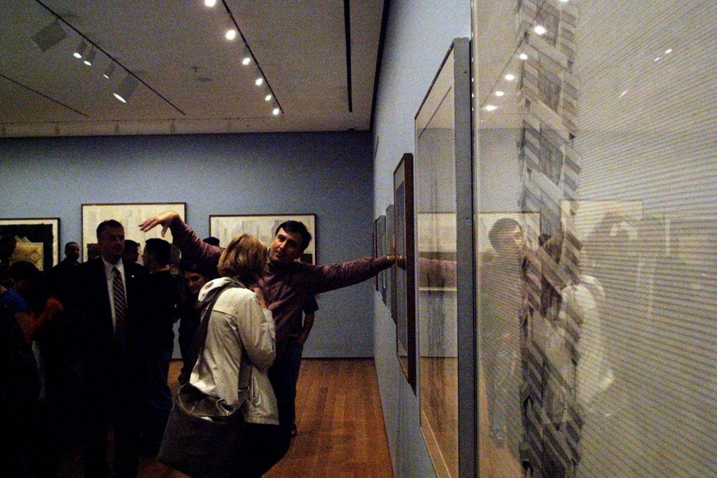 A group of people in a gallery, with one person gesturing toward framed artworks on the wall as others observe and discuss.