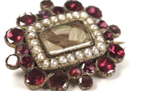 A close-up photograph of an ornate mourning brooch featuring a rectangular compartment at its center, containing intricately woven strands of a deceased relative’s hair. The compartment is framed by a border of small pearls, and the outer edge is adorned with deep red gemstones set in gold-toned metal. The brooch reflects the 19th-century tradition of memorial jewelry, preserving a tangible connection to lost loved ones.