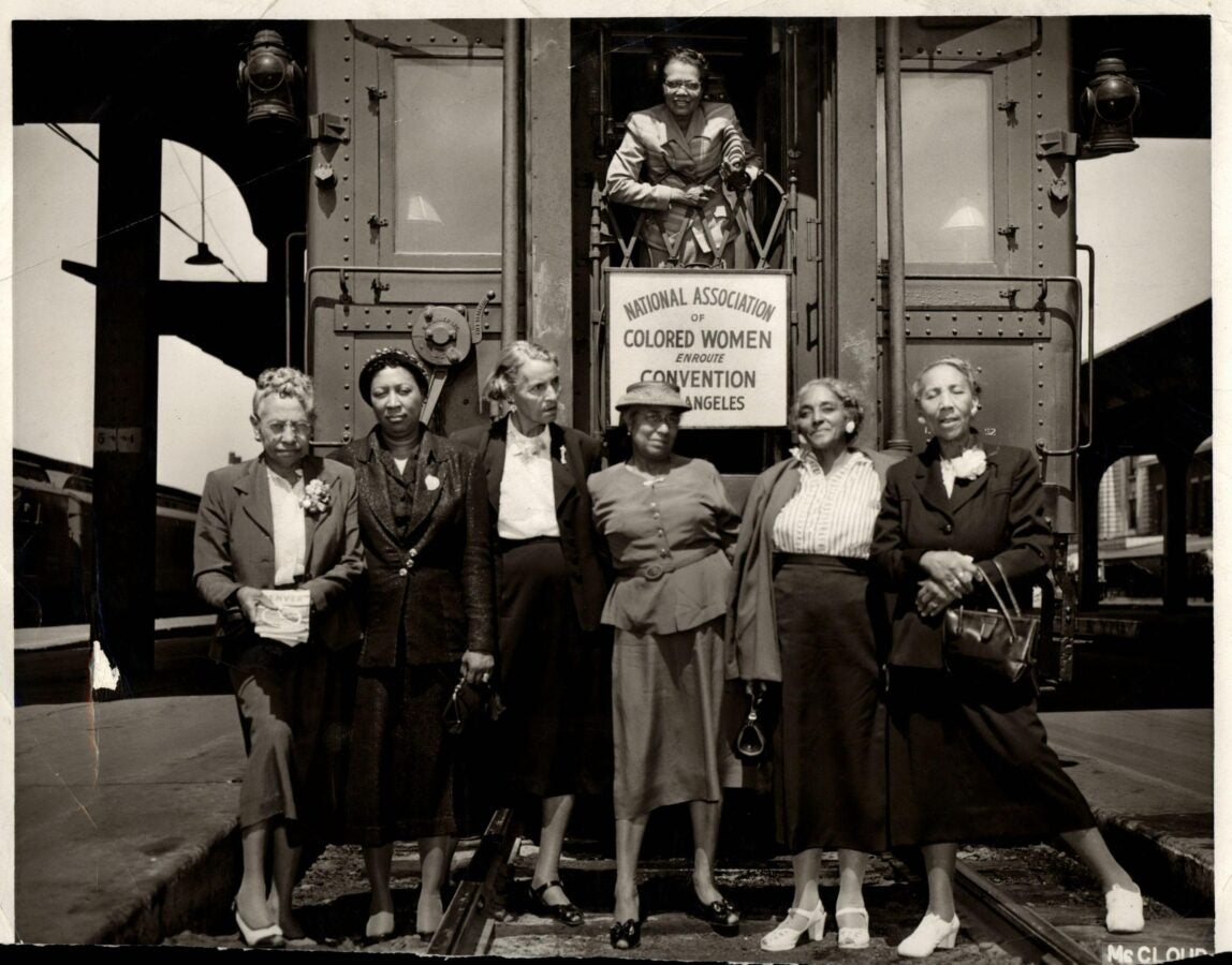 A black-and-white photograph of six African American women standing on a train platform in front of a railcar, with one woman leaning out of the train doorway. A sign on the train reads: “National Association of Colored Women Enroute Convention Los Angeles.” The women are dressed in formal attire, wearing suits, dresses, and hats, and carrying handbags. The image, taken between 1948-1952, captures a moment from the NACW’s journey to their convention.