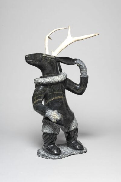 Stone and antler sculpture of a standing humanoid figure with the head of a caribou and full antlers. The figure wears detailed winter clothing, including mitts, boots, and a textured parka trim, and lifts one hand to its hood in a dynamic pose.