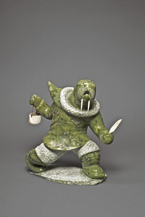 Green stone sculpture of a part-human, part-walrus figure in mid-motion, holding a knife in one hand and a pail in the other. The figure has a walrus head with ivory tusks, wearing a textured parka and standing on a stone base.