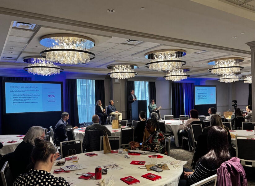 A conference room filled with round tables and attendees watching a panel discussion. Four speakers sit at the front, one standing at a podium, with presentation slides projected on either side of the stage. The slide highlights challenges in library workflows, including a statistic that 95% of collections are unprocessed. The room features elegant chandeliers and large windows with dark curtains.