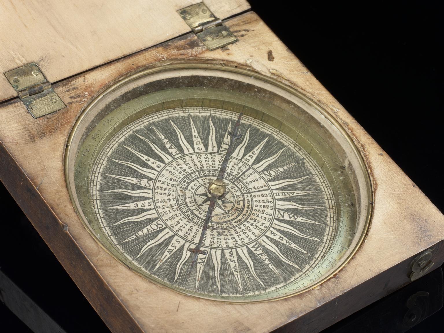 A 17th-century magnetic compass housed in a wooden case with brass hinges. The compass features a detailed wind rose with cardinal directions and degree markings engraved on a circular plate. The needle points north, and the ornate design includes concentric rings of numbers and letters used for navigation.