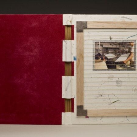 A uniquely bound book with a white, speckled handmade paper cover and red suede interior. The cover features a recessed window containing a photograph of someone working in a print or bindery studio. The binding uses exposed wooden elements and visible stitching.