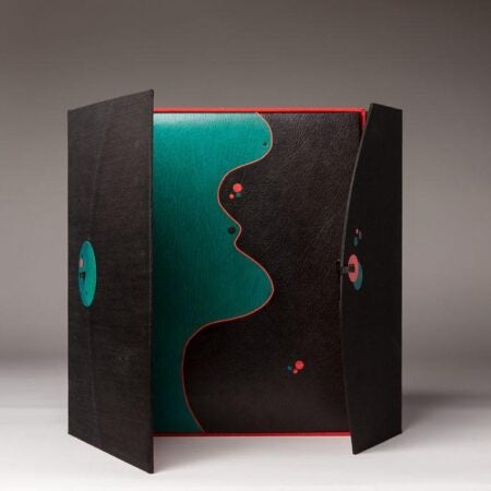 A sculptural book structure with two black doors partially opened to reveal a teal and black inner cover. The teal cover features a red profile silhouette of a face, with abstract circular embellishments. The dramatic contrast and geometric forms give the piece a modern, abstract feel.