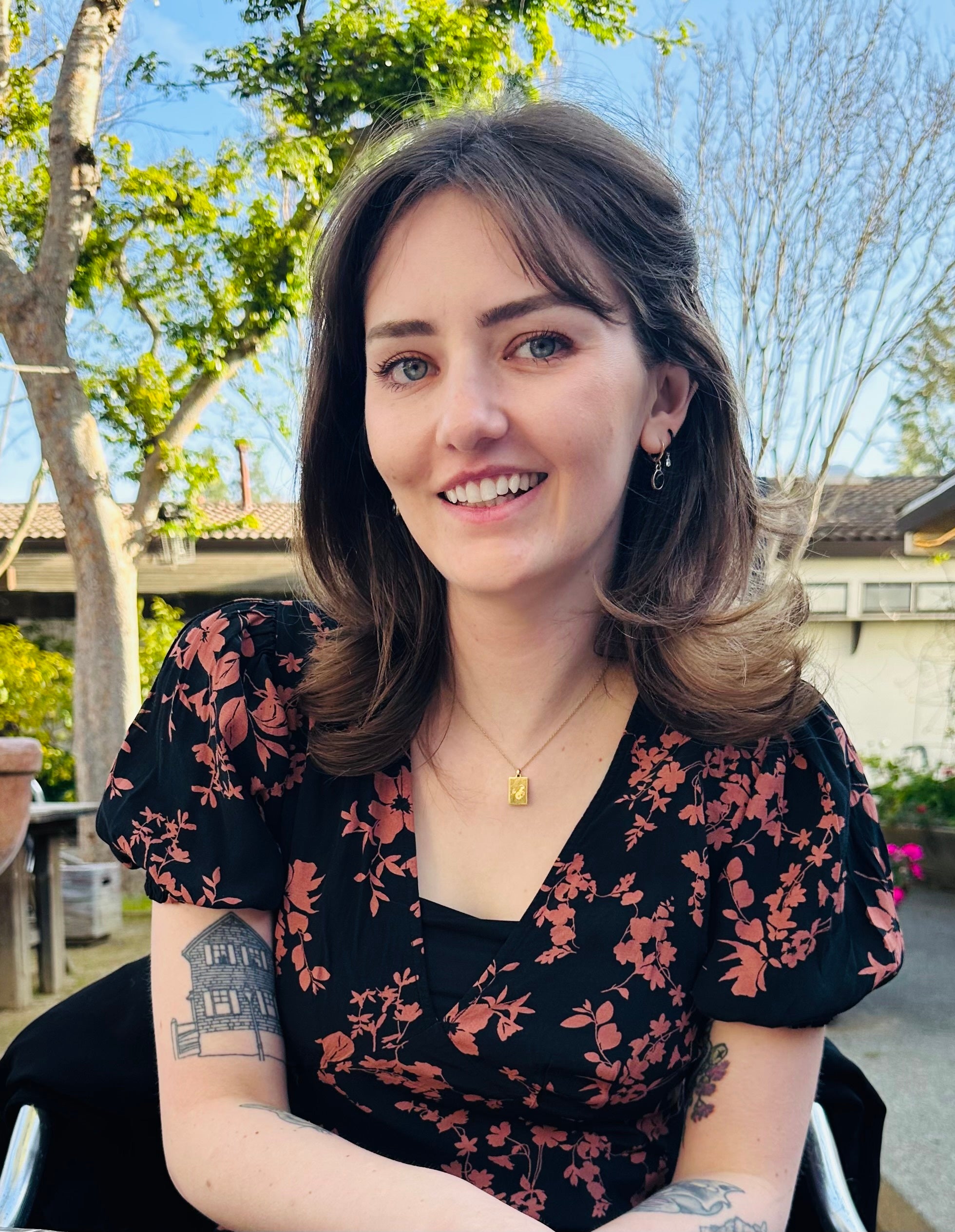 A woman with shoulder-length brown hair sits outside smiling. She is wearing a black dress with a red floral pattern, small hoop earrings, and a gold necklace. Trees and a house with a tiled roof are visible in the background.