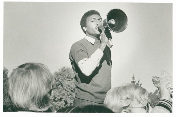 A black-and-white photograph of a man speaking passionately through a megaphone at an outdoor gathering. He wears a light shirt under a dark sweater and stands above a crowd, with trees visible in the background. Several people in the foreground, some with light-colored hair and glasses, listen closely and raise their hands.