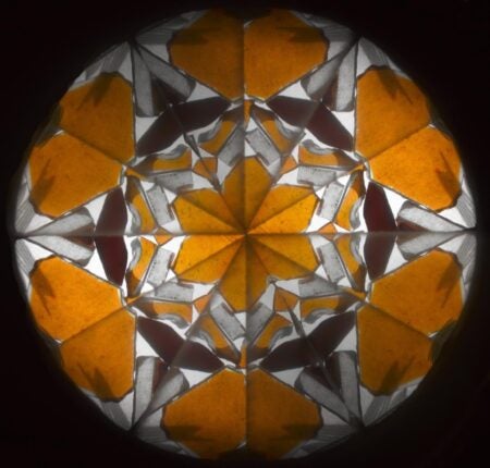 A kaleidoscope pattern viewed through Brewster’s Patent Kaleidoscope (1815–1825). The symmetrical design features triangular shapes in warm golden-yellow and white, forming a starburst effect with repeating geometric reflections on a dark circular background.