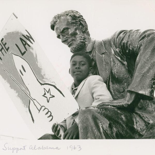 A young Black boy sits beside a large bronze statue of Abraham Lincoln, holding a protest sign with a drawing of a Ku Klux Klan hooded figure labeled “THE LAW.” The handwritten caption at the bottom reads “Support Alabama 1963.”