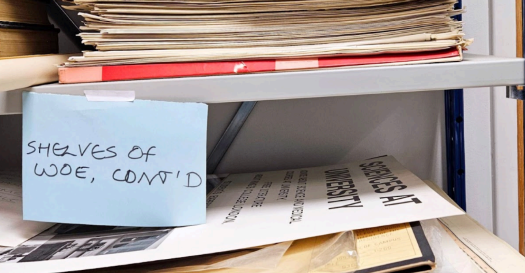 A cluttered metal shelf filled with stacks of old papers and folders. A blue sticky note taped to the shelf reads “SHELVES OF WOE, CONT’D” in black marker.
