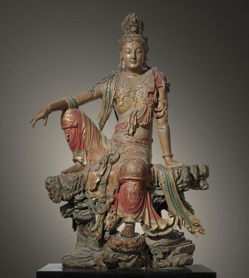 Painted and gilded wooden statue of Guanyin seated in a relaxed ‘royal ease’ pose on a rocky base, with flowing red-green robes and an elaborate crown against a neutral backdrop. (Chinese, Liao/Jin dynasty)