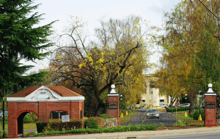 A brick entrance gate with a sign reading "Oregon State Penitentiary" is surrounded by trees with autumn leaves. The scene is set in a quiet area, with a building visible in the background.