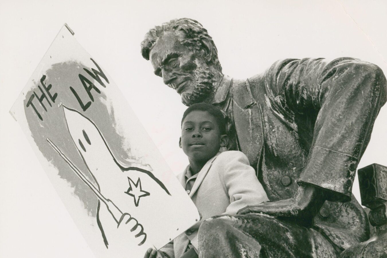 A young Black boy sits beside a statue of Abraham Lincoln, holding a protest sign reading “The Law” with a drawing of a Ku Klux Klan hooded figure. The handwritten caption below reads “Support Alabama 1963.”