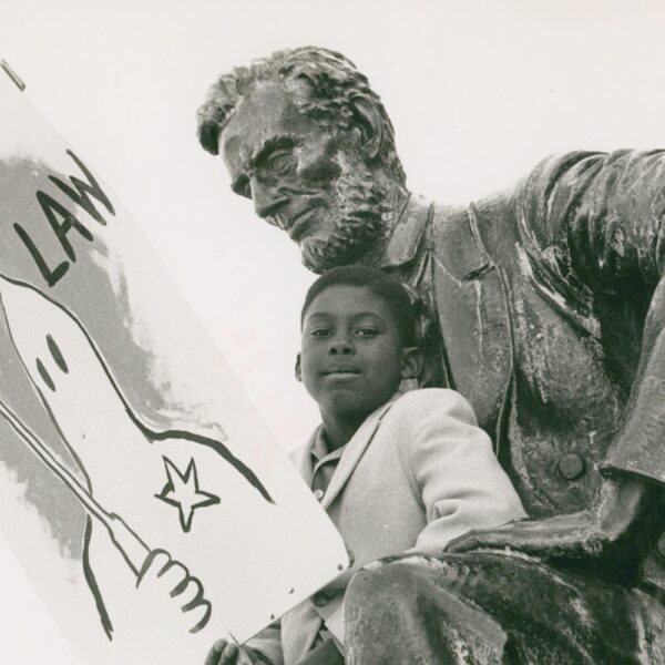A young Black boy sits beside a statue of Abraham Lincoln, holding a protest sign reading “The Law” with a drawing of a Ku Klux Klan hooded figure. The handwritten caption below reads “Support Alabama 1963.”