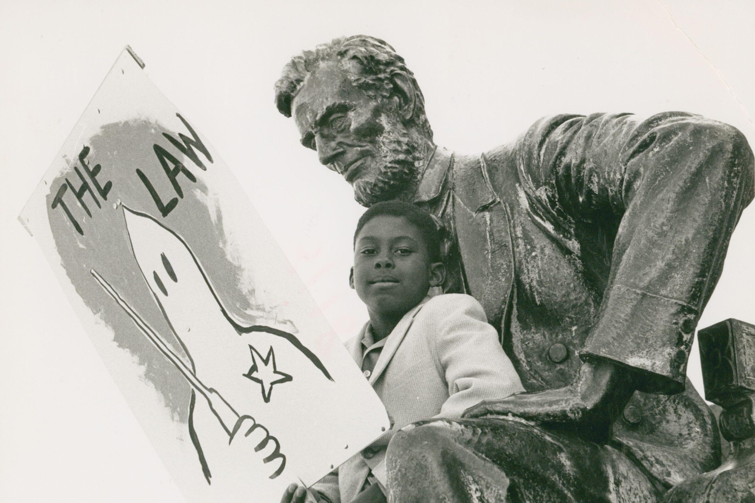 A young Black boy sits beside a statue of Abraham Lincoln, holding a protest sign reading “The Law” with a drawing of a Ku Klux Klan hooded figure. The handwritten caption below reads “Support Alabama 1963.”