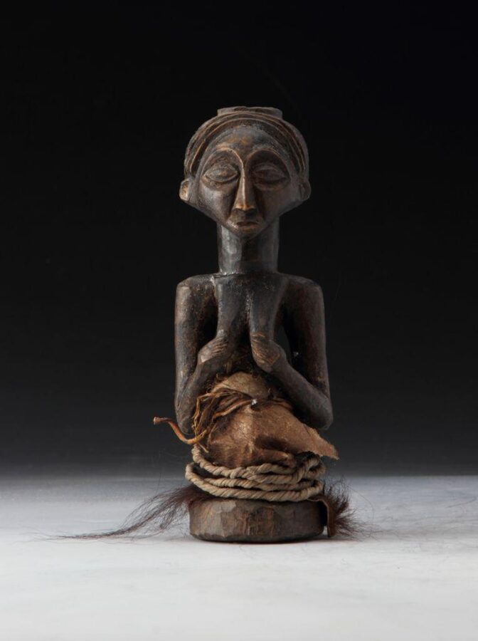 A carved wooden figure with stylized facial features, seated with hands clasped at the chest, wrapped in rope and fur material around the waist.