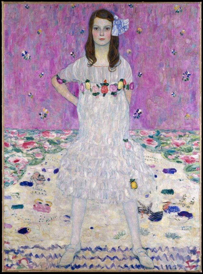 A portrait of a young girl standing confidently in a white ruffled dress decorated with colorful flowers, set against a bright pink patterned background. Painted in an expressive, textured style by Gustav Klimt.