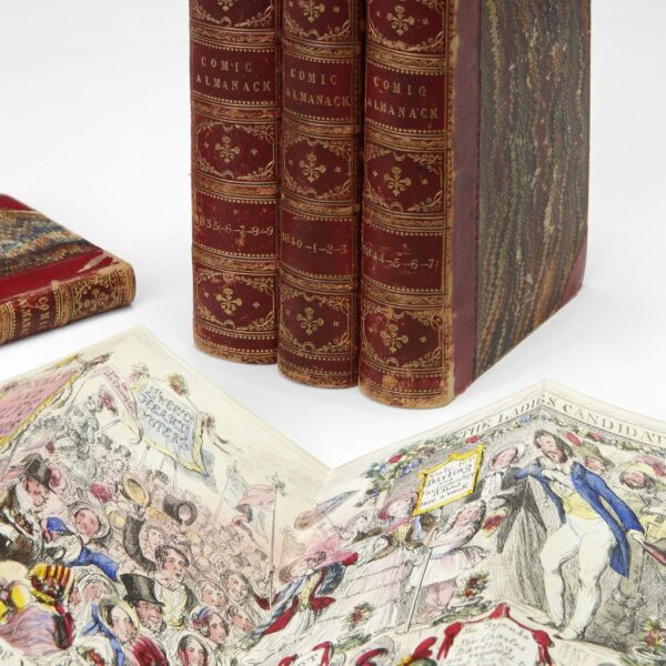 Three bound volumes of The Comic Almanack with red leather spines and marbled covers stand upright beside an open book showing a colorful illustrated page. The page depicts a lively 19th-century crowd scene with banners and caricatured figures, featuring ornate period costumes and detailed linework.