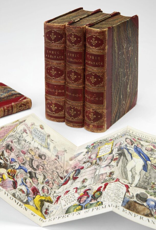 Three bound volumes of The Comic Almanack with red and gold spines, shown alongside an open illustrated page featuring a colorful satirical scene.