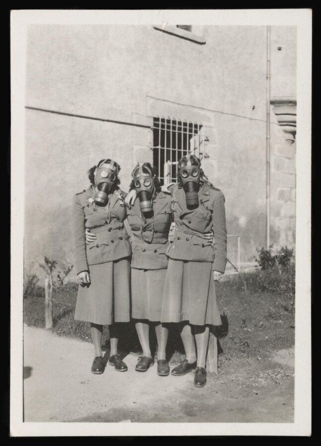 Black-and-white photo of three women in military uniforms wearing gas masks, standing arm in arm outdoors.