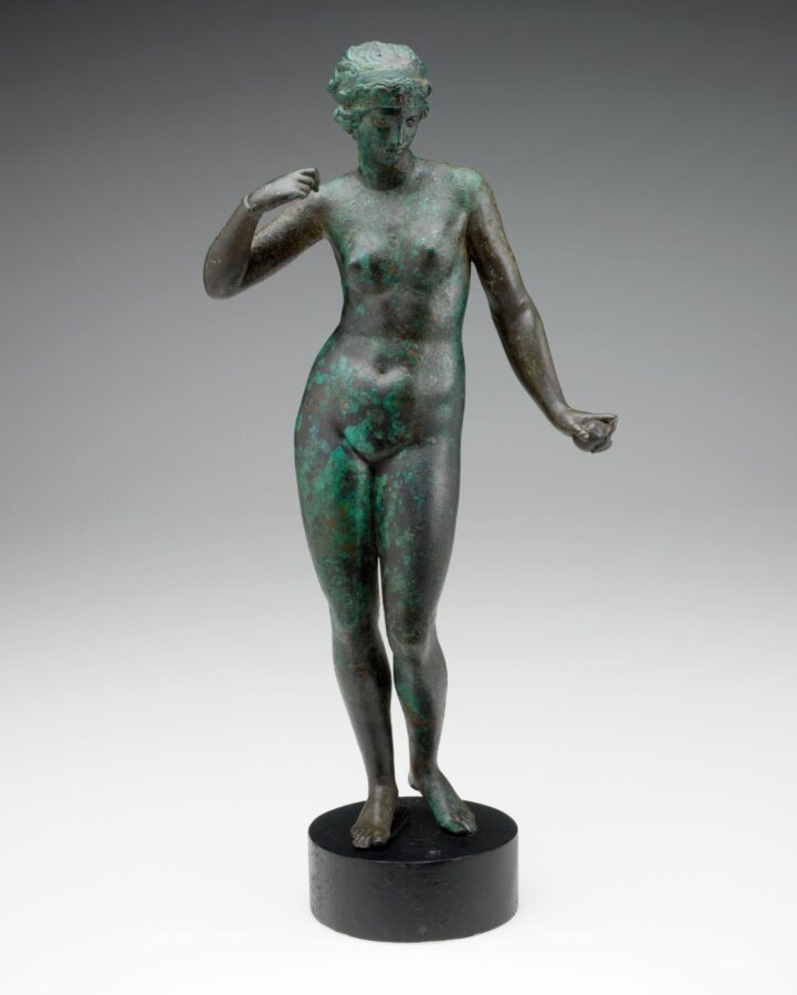 A bronze statue of Aphrodite from 2nd century BCE Greece, depicting a nude female figure with a slightly turned head and raised arm, standing on a round black base.