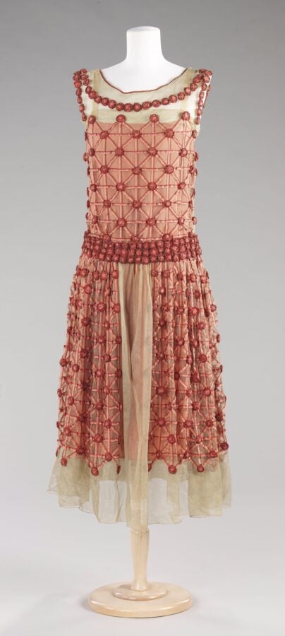 Sleeveless 1920s evening dress with a geometric lattice of red rosettes and embroidery over sheer beige fabric, displayed on a mannequin.