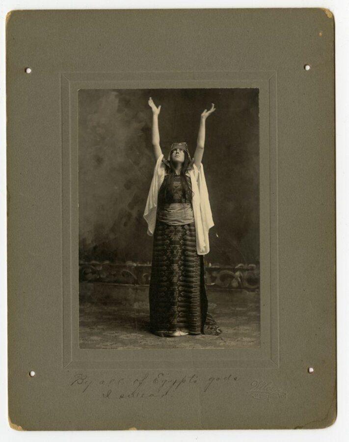 Studio portrait of a woman in a long patterned dress and headpiece raising her arms in a dramatic pose, inscribed “By all of Egypt’s gods I swear!”
