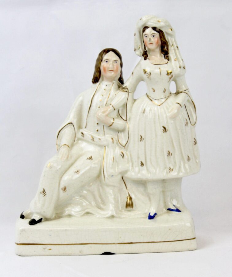 A white ceramic figurine depicting a man seated beside a standing woman in 19th-century attire, detailed with gold accents and blue shoes.