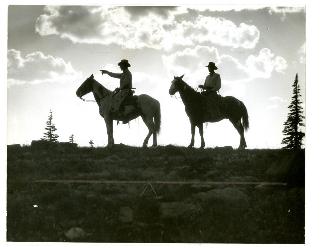 Silhouette of two cowboys on horseback atop a ridge in Wyoming, one pointing into the distance against a backdrop of clouds and open sky.