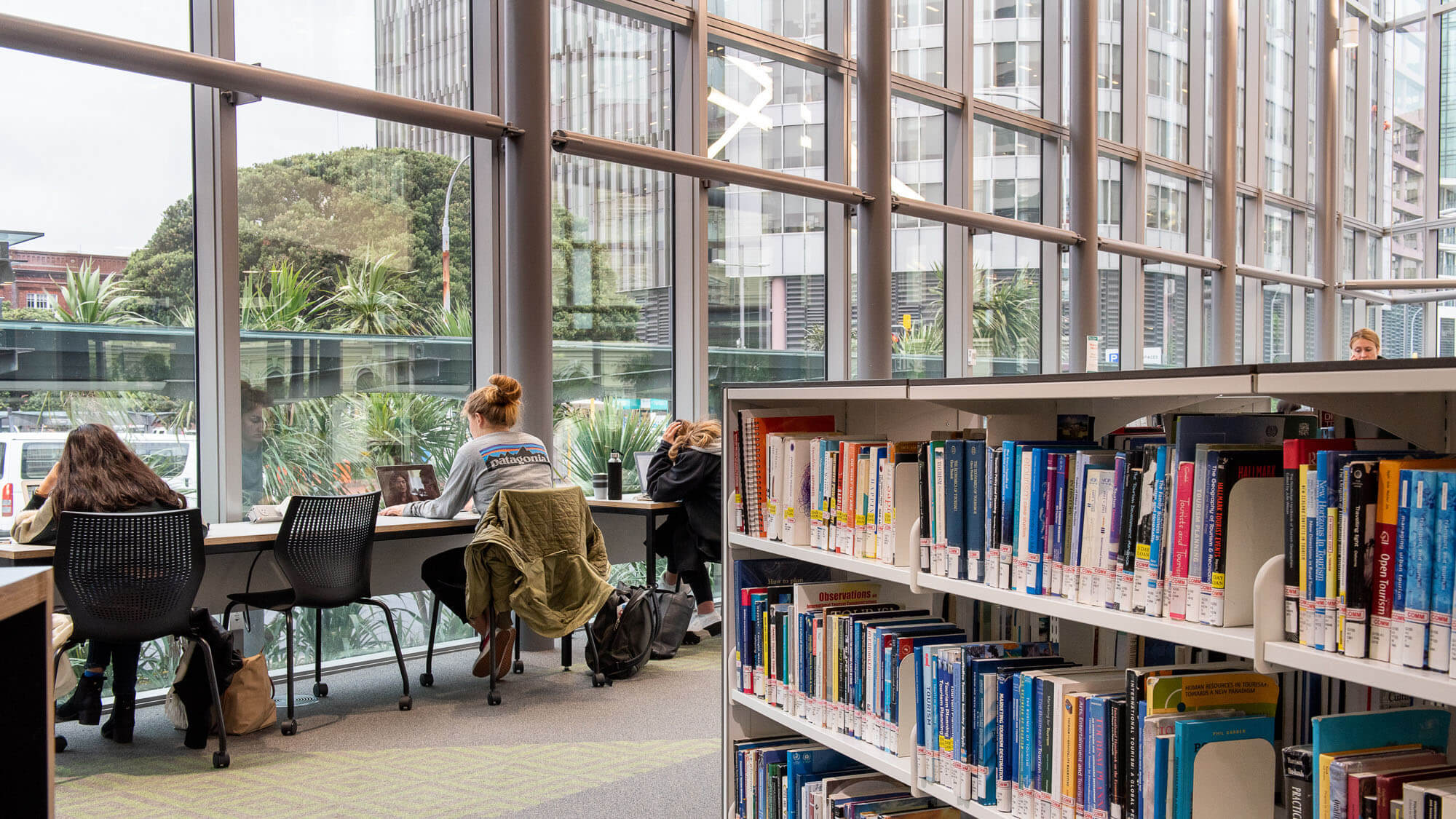 Students study at desks beside large windows overlooking greenery inside the library at Victoria University of Wellington.