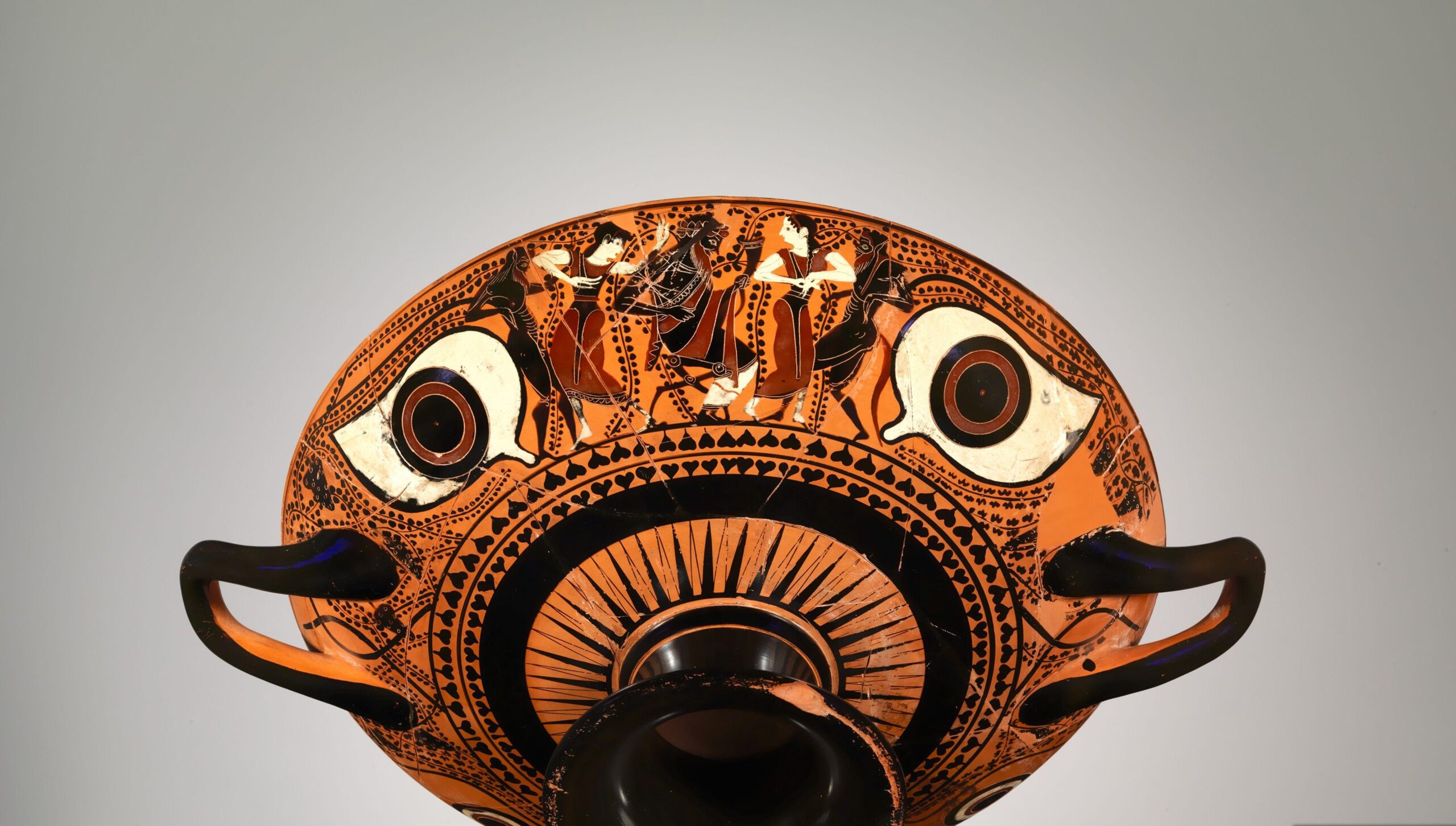 A terracotta kylix eye cup from around 530 BCE featuring large painted eyes and a black-figure scene of dancing figures across the top band on an orange background.