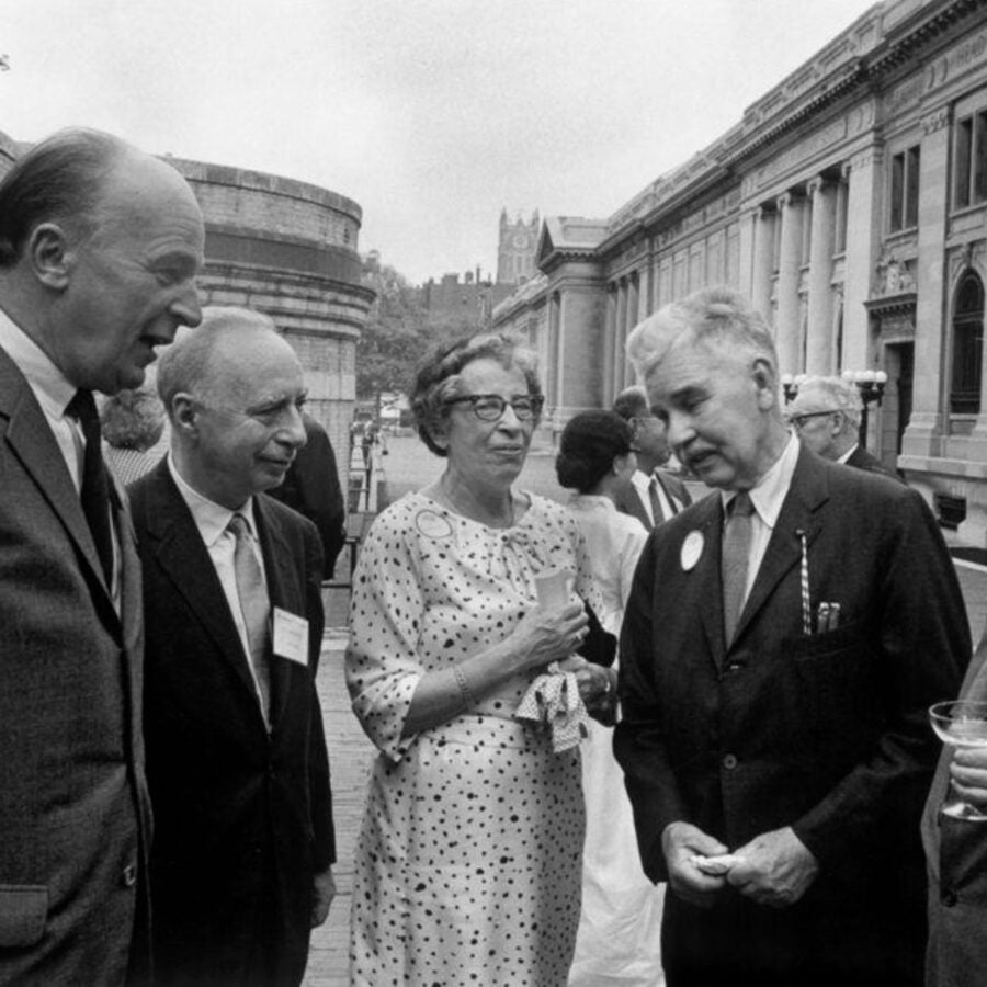Black-and-white photograph of a group of five people in formal attire with Hannah Arendt at the center, conversing outdoors near a large building, possibly at an academic or professional event.