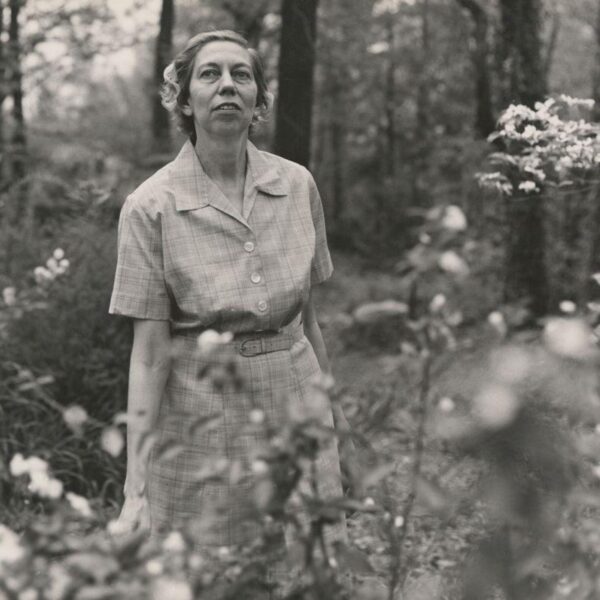 Black-and-white photograph of Eudora Welty standing outdoors among flowering plants, wearing a short-sleeved checked dress.