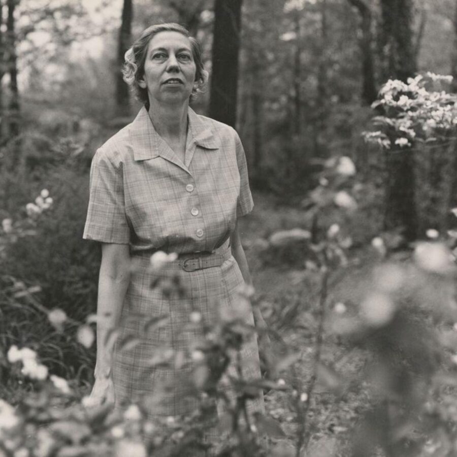 Black-and-white photograph of Eudora Welty standing outdoors among flowering plants, wearing a short-sleeved checked dress.