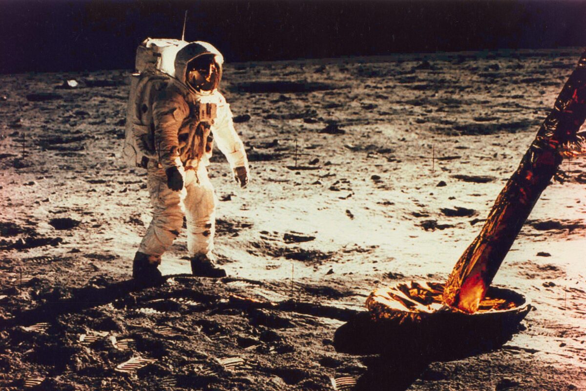Astronaut in a white spacesuit standing on the moon’s surface near a lander footpad, with footprints and lunar soil illuminated against the dark sky.