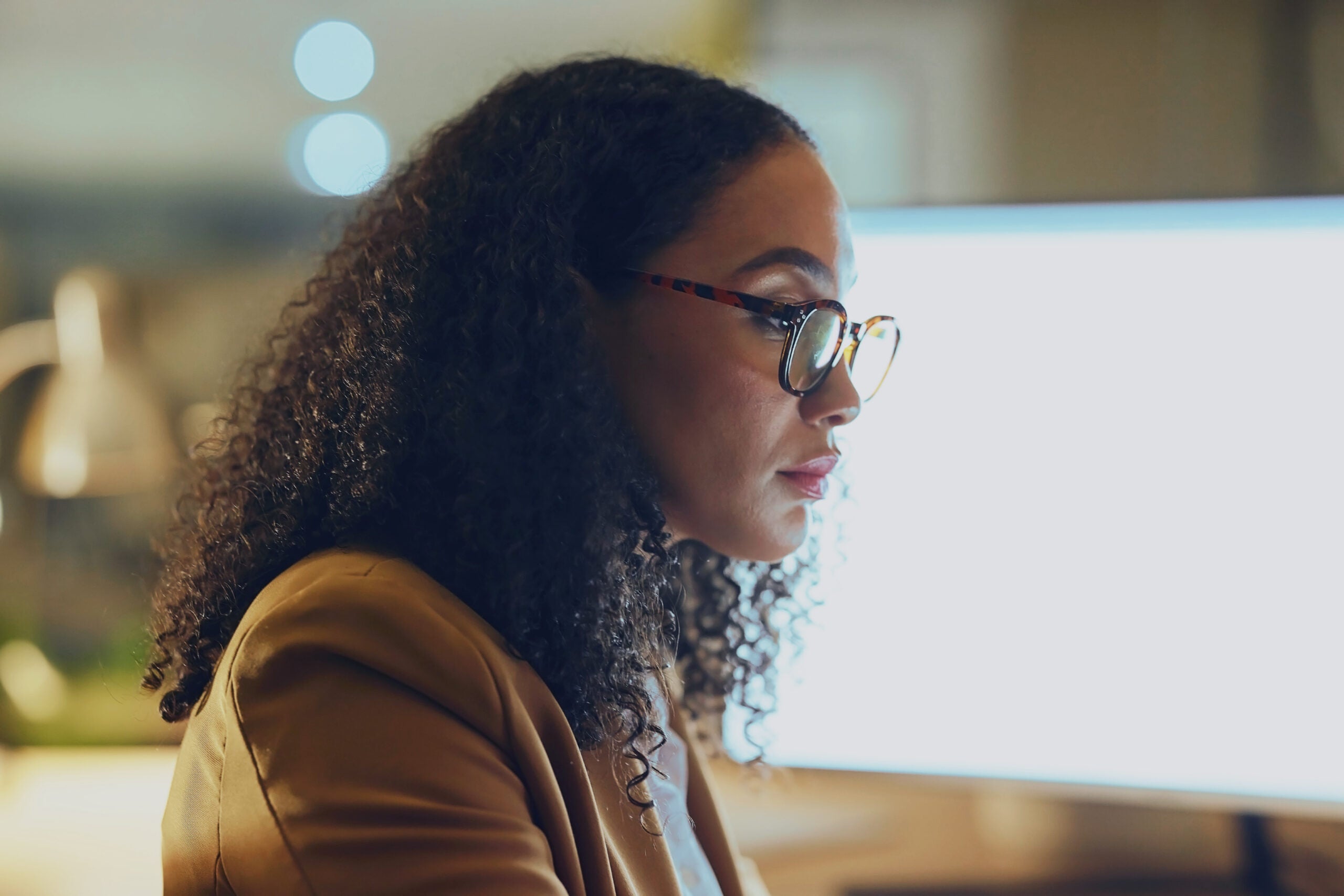 A woman with curly hair and glasses, wearing a tan blazer, works intently at a computer in a softly lit office. The monitor glows brightly in front of her as she focuses on the screen.