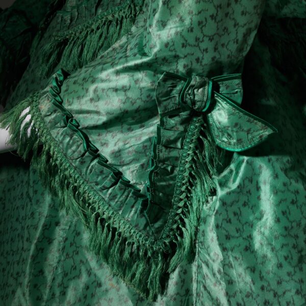 Close-up of a 19th-century green silk dress with patterned fabric, fringe, ruffles, and a bow detail on the sleeve.