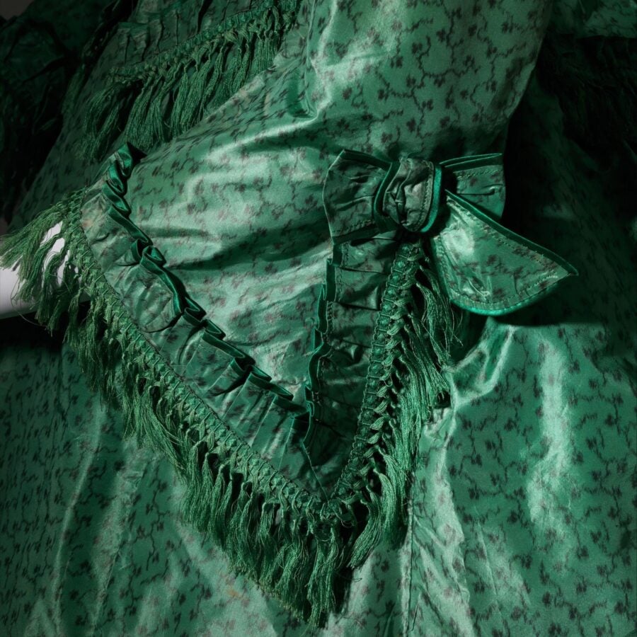 Close-up of a 19th-century green silk dress with patterned fabric, fringe, ruffles, and a bow detail on the sleeve.