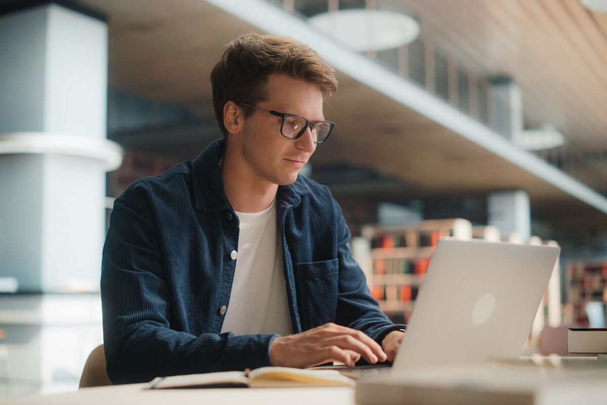 A student wearing glasses works on a laptop in a bright library setting.