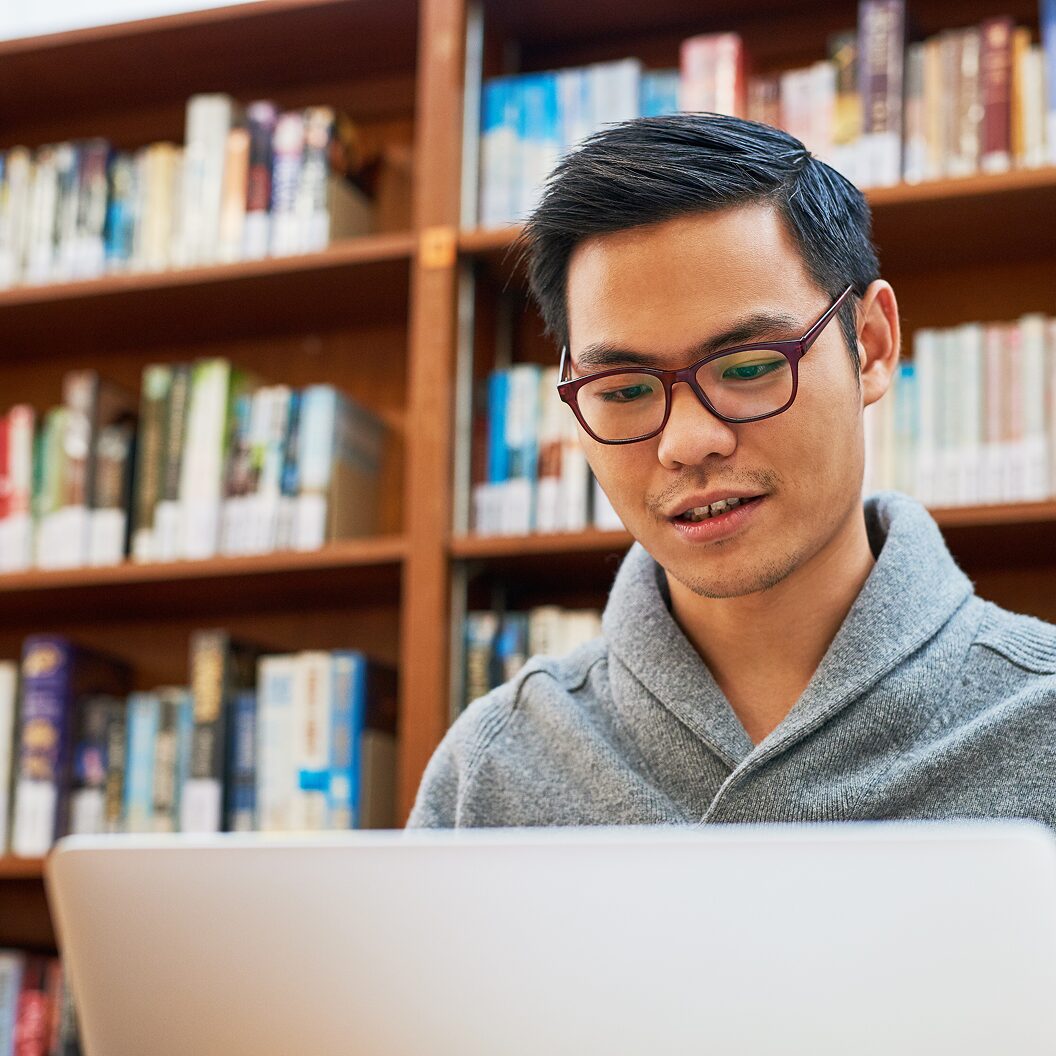 A person wearing glasses works on a laptop in a library, surrounded by bookshelves.
