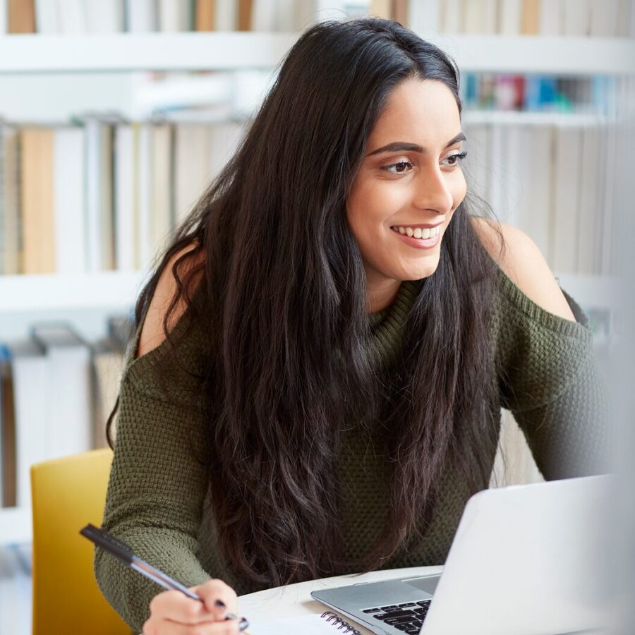 Person smiling while taking notes beside a laptop in a library with bookshelves behind them.