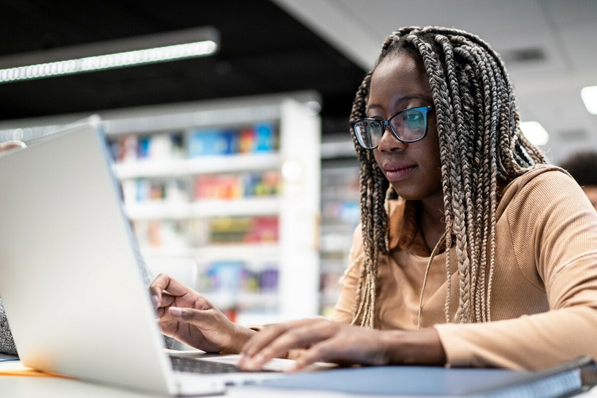 Person typing on a laptop in a library or study area with shelves of books in the background.