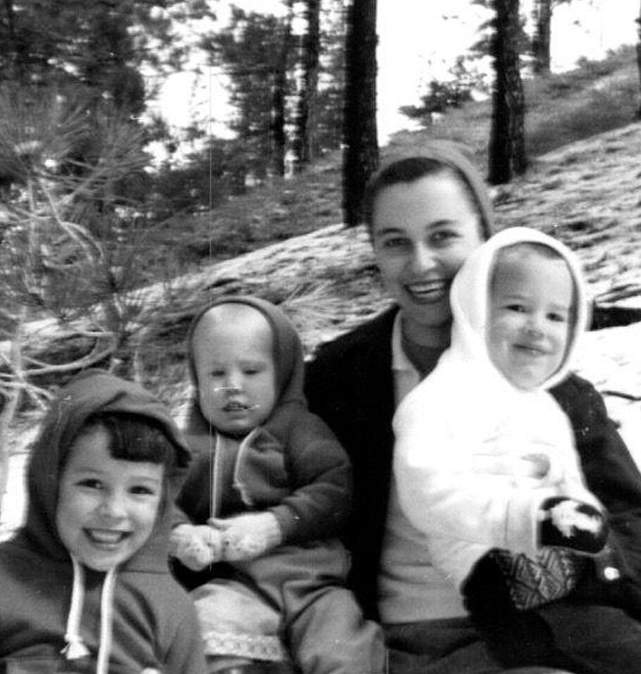 Black-and-white photo of a smiling woman outdoors with three young children dressed in hooded winter clothing.