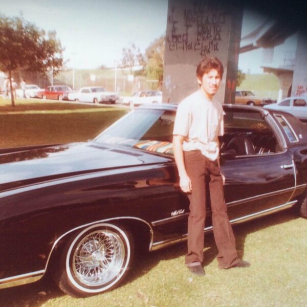 Color photo from the 1970s of a young man standing beside a classic black car with chrome wheels in a sunny park.