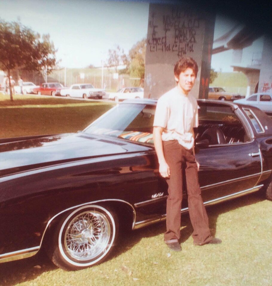 Color photo from the 1970s of a young man standing beside a classic black car with chrome wheels in a sunny park.