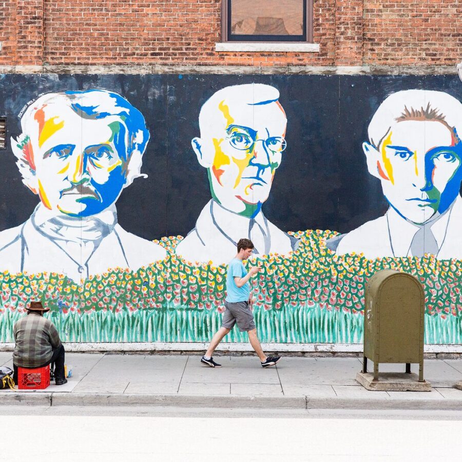 Colorful mural on a brick wall featuring stylized portraits of three historical figures, with two people visible on the sidewalk below.