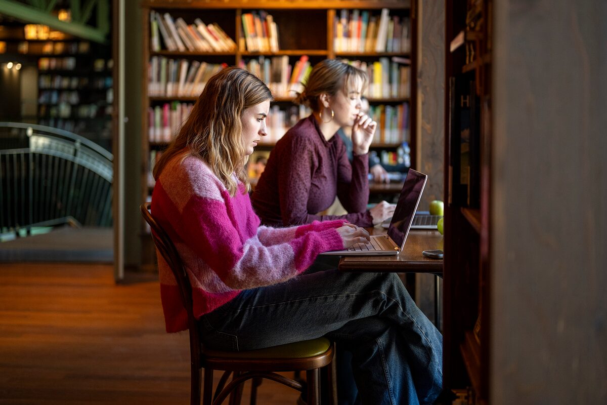 Two people work on laptops at a wooden table in a library, focused on their screens with bookshelves filled with colorful books in the background.